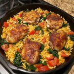 Skillet of seasoned chicken thighs cooked with orzo pasta, spinach, and tomatoes.