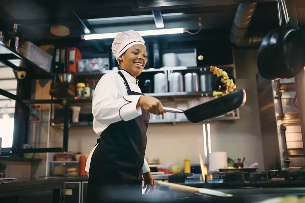 A smiling chef tossing vegetables in a pan in a commercial kitchen.