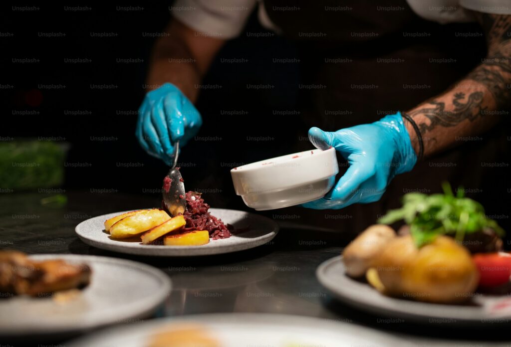 Chef wearing gloves and carefully plating a dish with sanitized tools for hygienic presentation in a restaurant kitchen.